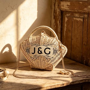 May include: Woven handbag with a handle and a gold chain strap. The bag is beige with the letters "J&G" in black, framed by floral and leaf designs. The bag sits on a wooden surface.