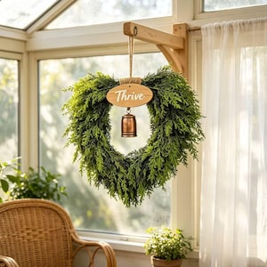 May include: A heart-shaped wreath made of green faux cedar foliage hanging from a wooden wall bracket. An oval wooden plaque with the word Thrive in white script is attached to the top, and a small copper-coloured metal bell hangs below it.