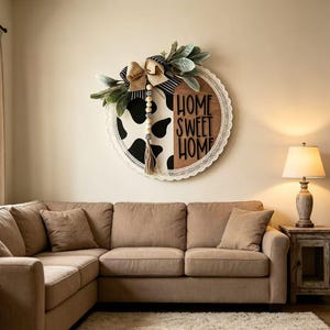 May include: A circular wall sign featuring a split design with a black and white cow print on one side and a natural wood finish on the other. The text HOME SWEET HOME is in black lettering. It is decorated with a burlap bow, greenery, and beads.