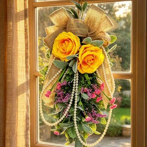 May include: A decorative floral swag featuring two large yellow roses, green foliage, and small purple flowers. The arrangement is accented with a large tan burlap bow at the top and draped with long strands of white faux pearls hanging downwards.