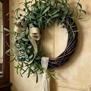 May include: A circular wreath constructed from dark brown woven branches, adorned with green olive branches featuring small black olives. A beige ribbon is tied in a bow on the left side, and a long, cream-coloured rope tassel hangs from the bottom.