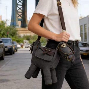 Peut inclure: Un sac bandoulière en toile grise en forme de cheval. Le sac est orné d'une selle et d'une sangle en cuir marron, avec des boucles et des chaînes dorées. La tête du cheval présente un bridon détaillé. Le sac est porté à l'épaule.