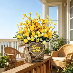 May include: A floral arrangement featuring bright yellow forsythia branches and yellow tulips in a rectangular woven basket. A heart-shaped wooden sign with the text Love wins. is attached to the front, accented by a tan burlap ribbon bow.