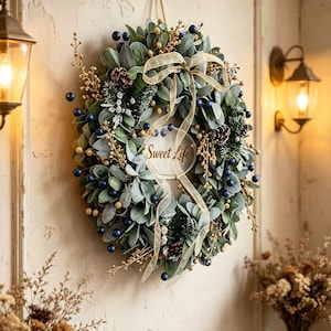 Peut inclure: Une couronne circulaire composée de feuilles vertes, de baies bleu foncé, de brindilles dorées et de pommes de pin. Un ruban doré transparent est noué au sommet, et un panneau circulaire central affiche le texte Sweet Life en écriture cursive.