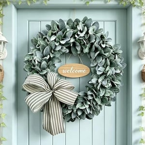 May include: A circular wreath made of sage green lamb's ear leaves, featuring a large black and white striped fabric bow at the bottom and a centered oval wooden sign with the word "welcome" printed in white script lettering.