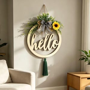 May include: A circular wooden wall sign featuring the word hello in cursive script. The top is decorated with green foliage, a yellow sunflower, and a black and white checkered bow. A dark green tassel hangs from the bottom of the wooden ring.
