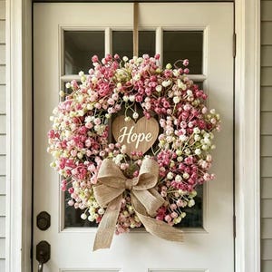 以下が含まれることがあります： 小さなピンクと白の花で作られた円形のリースで、中央には白い文字でHopeと書かれた木製のハート型のオーナメントが付いています。花飾りの下部には、質感のあるベージュの麻布で作られた大きなリボンが結ばれています。