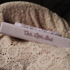 Puede incluir: Un libro cubierto de tela rosa claro con el texto Our Little Bud bordado en hilo rosa oscuro en el lomo. El libro presenta un borde de encaje blanco decorativo a lo largo del borde superior y tiene una apariencia suave y texturizada.