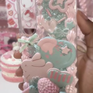 May include: A clear phone case decorated with pink and mint green faux whipped cream, featuring a pink rabbit figure, a mint green heart, a textured pink strawberry, a small green ribbon bow, and various star-shaped embellishments.