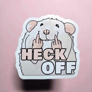 May include: A white sticker featuring a cartoon rat giving the middle finger with the words "HECK OFF" in pink and white block letters. The rat has pink ears and a pink nose, set against a light pink background.