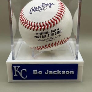 Op de afbeelding: Een honkbal in een doorzichtige vitrine. De bal is wit met rode stiksels en het Rawlings-logo. De tekst op de bal luidt "2021 ALL-STAR GAME". De basis van de behuizing is wit met een blauw bordje met de tekst "KC Bo Jackson".