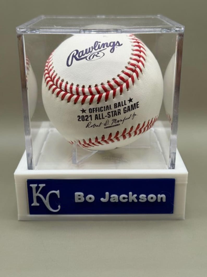 Op de afbeelding: Een honkbal in een doorzichtige vitrine. De bal is wit met rode stiksels en het Rawlings-logo. De tekst op de bal luidt "2021 ALL-STAR GAME". De basis van de behuizing is wit met een blauw bordje met de tekst "KC Bo Jackson".