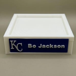 Op de afbeelding: Een witte rechthoekige bak met een opstaande rand. Een blauw rechthoekig label op de voorkant toont de tekst "KC" en "Bo Jackson" in witte letters. De bak is waarschijnlijk van plastic.