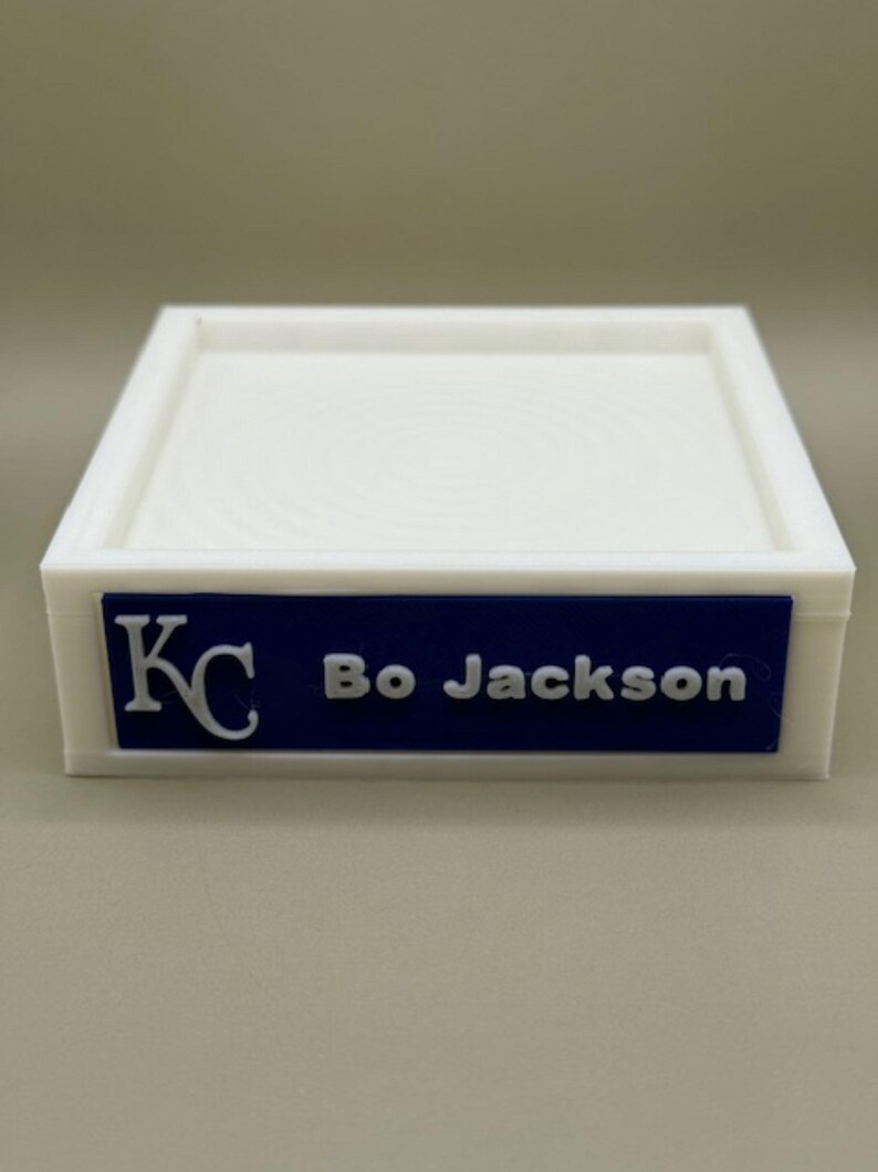 Op de afbeelding: Een witte rechthoekige bak met een opstaande rand. Een blauw rechthoekig label op de voorkant toont de tekst "KC" en "Bo Jackson" in witte letters. De bak is waarschijnlijk van plastic.