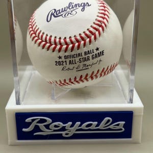 May include: A white baseball with red stitching, displayed in a clear case. The ball is marked "Rawlings" and "2021 All-Star Game." The base of the case features the word "Royals" in white on a blue background.