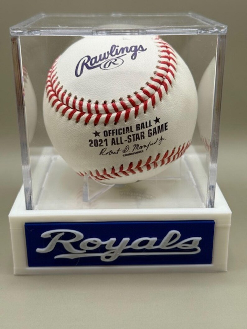 May include: A white baseball with red stitching, displayed in a clear case. The ball is marked "Rawlings" and "2021 All-Star Game." The base of the case features the word "Royals" in white on a blue background.