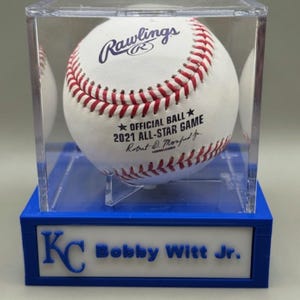 Op de afbeelding: Een witte honkbal met rode stiksels, tentoongesteld in een heldere acryl behuizing op een blauwe basis. De bal is gemarkeerd met "Rawlings" en "OFFICIAL BALL 2021 ALL-STAR GAME". De basis leest "KC Bobby Witt Jr."