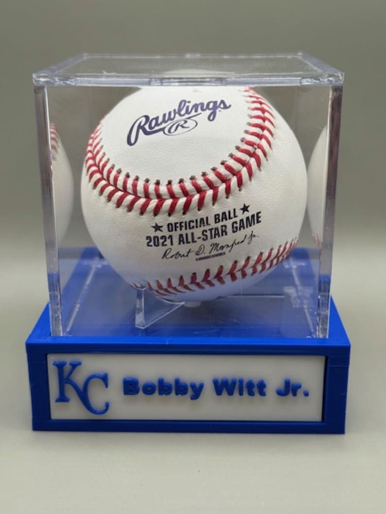 Op de afbeelding: Een witte honkbal met rode stiksels, tentoongesteld in een heldere acryl behuizing op een blauwe basis. De bal is gemarkeerd met "Rawlings" en "OFFICIAL BALL 2021 ALL-STAR GAME". De basis leest "KC Bobby Witt Jr."