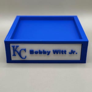 Op de afbeelding: Een blauwe rechthoekige lade met een wit paneel met de letters "KC" en de naam "Bobby Witt Jr." in blauw. De lade heeft een verhoogde rand en een plat interieur.