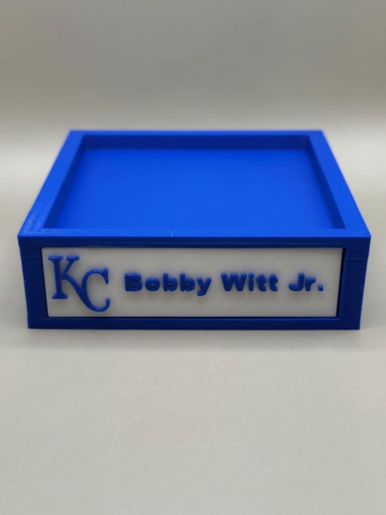 Op de afbeelding: Een blauwe rechthoekige lade met een wit paneel met de letters "KC" en de naam "Bobby Witt Jr." in blauw. De lade heeft een verhoogde rand en een plat interieur.