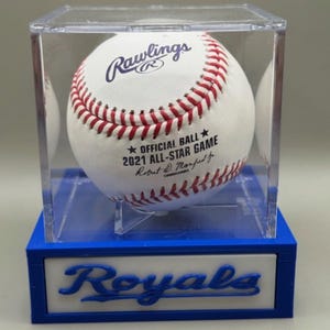 May include: A white Rawlings baseball with red stitching, displayed in a clear acrylic case. The ball is labeled "2021 All-Star Game" and is set on a blue and white base with "Royals" text.