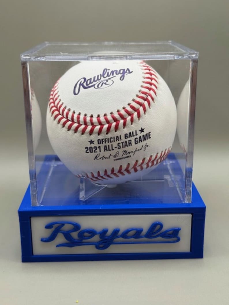 May include: A white Rawlings baseball with red stitching, displayed in a clear acrylic case. The ball is labeled "2021 All-Star Game" and is set on a blue and white base with "Royals" text.