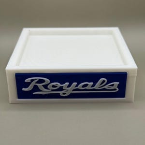 May include: A white, square tray with a raised edge. A blue rectangular accent features the word "Royals" in white script. The tray is likely designed for small items or display.