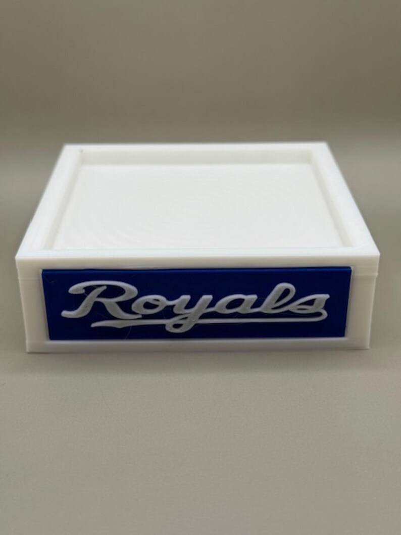 May include: A white, square tray with a raised edge. A blue rectangular accent features the word "Royals" in white script. The tray is likely designed for small items or display.