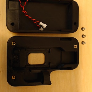 May include: Black, open-top, rectangular plastic case with a compartment and a smaller section. Red and black twisted wires with white connectors are visible inside. Three small screws are to the right. The case is on a light brown surface.