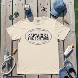 Op de afbeelding: Een crèmekleurig T-shirt met de tekst "CAPTAIN OF THE PONTOON" in een marineblauw ovaal ontwerp. Het shirt is omgeven door een zonnebril, een hoed, sleutels, een blikje frisdrank, zonnebrandcrème en blauwe sandalen, allemaal op een houten steiger.
