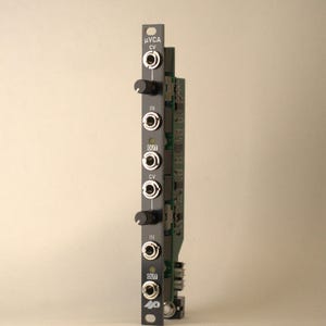 May include: A black modular synthesizer VCA module with a vertical faceplate featuring the text µVCA, CV, IN, and OUT. The unit includes two black rotary knobs, six silver input and output jacks, and a green circuit board visible on the side.