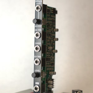 May include: A black electronic module with silver jacks, knobs, and the text "HH606 CV/GT". The module has several ports labeled "Accent", "OH TRIG", "Choke", "CH TRIG", "Level", and "OUT". Additional electronic components are visible.