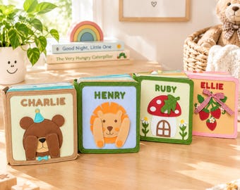 Puede incluir: Cuatro libros de tela cuadrados con aplicaciones de fieltro. El primero muestra un oso marrón con el nombre CHARLIE, el segundo un león con HENRY, el tercero una casa de setas con RUBY y el cuarto fresas con el nombre ELLIE.
