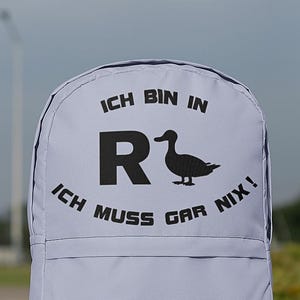 May include: A light gray backpack with black text and a duck silhouette. The text reads "ICH BIN IN R" above the duck and "ICH MUSS GAR NIX!" below. The backpack is outdoors.