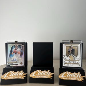 May include: Three black display stands with sports trading cards. The cards feature Drew Brees and Tyler Shough. Each stand has a gold and white "Clutch Collections AU" logo.