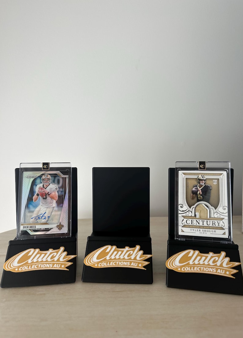May include: Three black display stands with sports trading cards. The cards feature Drew Brees and Tyler Shough. Each stand has a gold and white "Clutch Collections AU" logo.