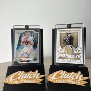 May include: Two football trading cards in protective cases on black display stands. One card features Drew Brees' signature. The stands have the text "Clutch Collections AU" in gold and white.