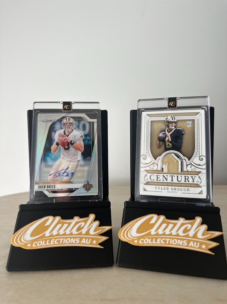 May include: Two football trading cards in protective cases on black display stands. One card features Drew Brees' signature. The stands have the text "Clutch Collections AU" in gold and white.