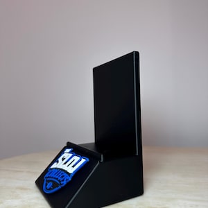 May include: A black display stand with a slanted base and a vertical back. A blue and white logo that says "SDI Collects" is affixed to the base. The stand is designed to hold and display items.