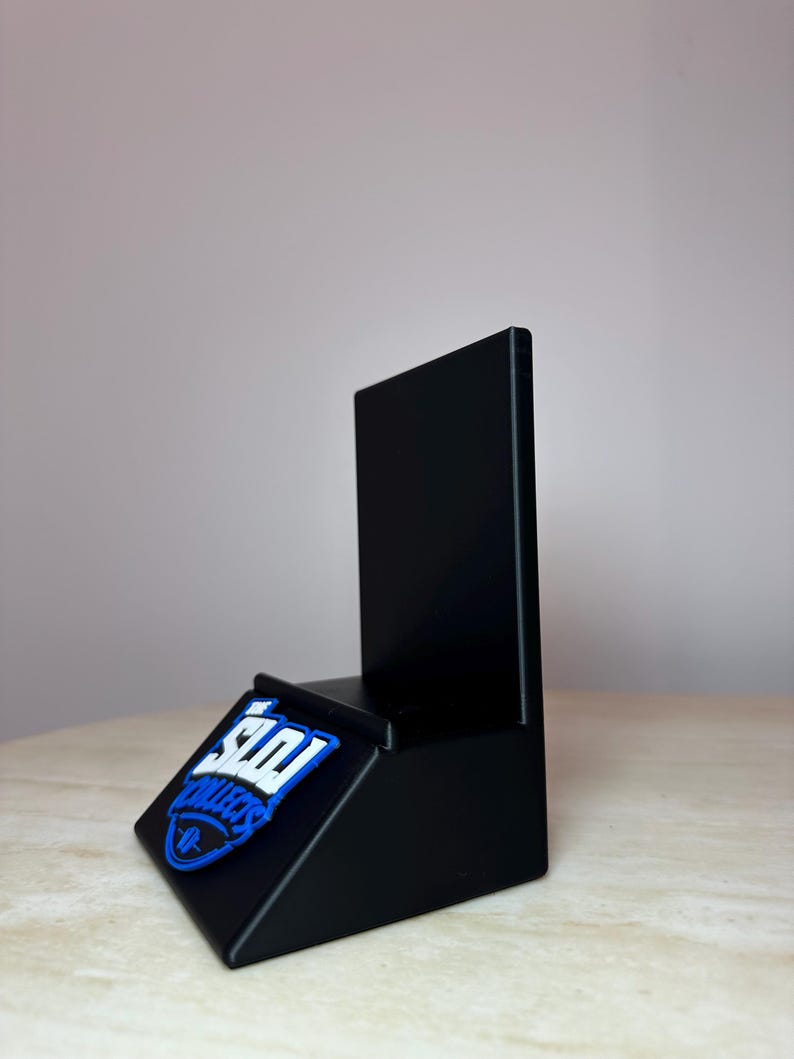 May include: A black display stand with a slanted base and a vertical back. A blue and white logo that says "SDI Collects" is affixed to the base. The stand is designed to hold and display items.