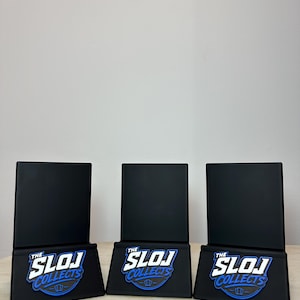 May include: Three black display stands with a rectangular top and a base featuring the text "THE SLOJ COLLECTS" in white and blue. The stands are arranged on a light brown surface against a plain white background.