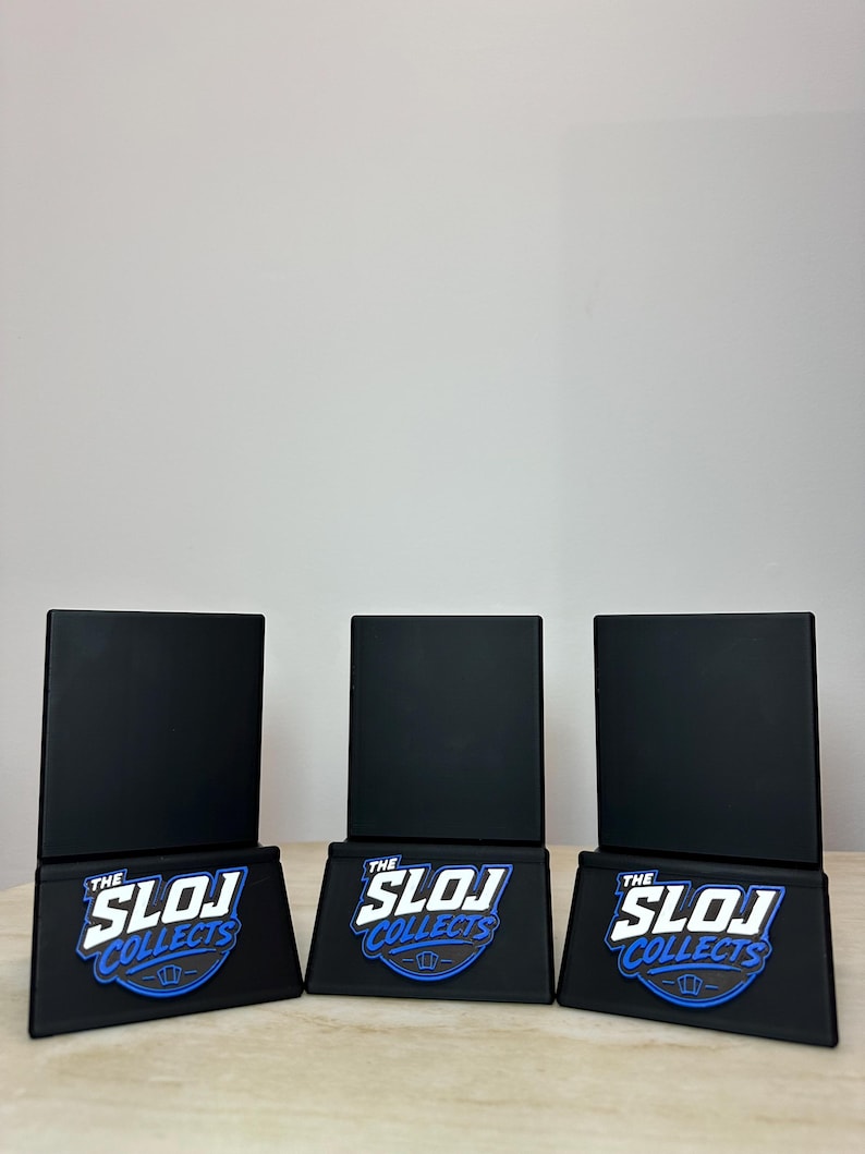 May include: Three black display stands with a rectangular top and a base featuring the text "THE SLOJ COLLECTS" in white and blue. The stands are arranged on a light brown surface against a plain white background.