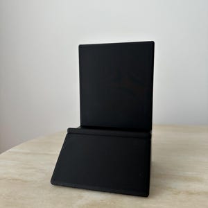 May include: A black, angled display stand. The stand has a rectangular base and a vertical back, suitable for holding items. The stand is made of a matte black material and sits on a light-colored surface.