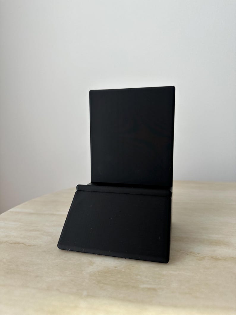 May include: A black, angled display stand. The stand has a rectangular base and a vertical back, suitable for holding items. The stand is made of a matte black material and sits on a light-colored surface.