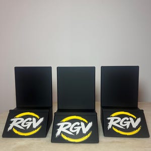 May include: Three black display stands with a rectangular back and a slanted base. Each base features the white letters "RGV" inside a yellow circle with a white outline. The stands are arranged in a row on a light-colored surface.