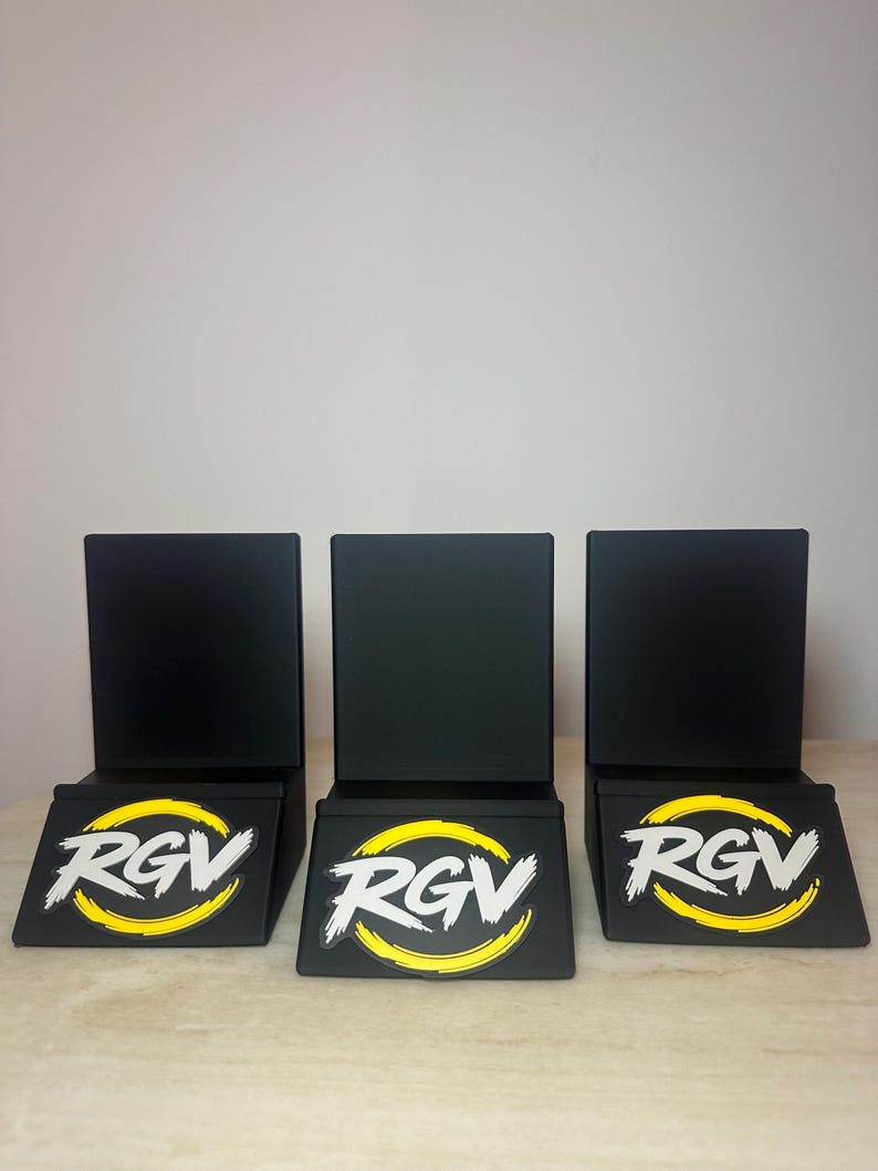May include: Three black display stands with a rectangular back and a slanted base. Each base features the white letters "RGV" inside a yellow circle with a white outline. The stands are arranged in a row on a light-colored surface.