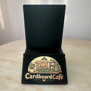 May include: Black tabletop phone stand with a decorative front panel. The panel features a cartoon illustration of a box character with the text "Cardboard Caf&eacute;" and the words "Buy Sell Trade".