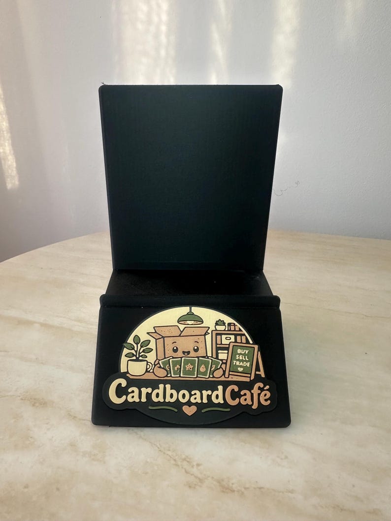 May include: Black tabletop phone stand with a decorative front panel. The panel features a cartoon illustration of a box character with the text "Cardboard Caf&eacute;" and the words "Buy Sell Trade".