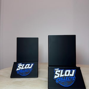 May include: Two black display stands with the text "THE SLOJ COLLECTS" in white and blue. The stands have a rectangular base and a square top. The stands are on a light-colored surface.