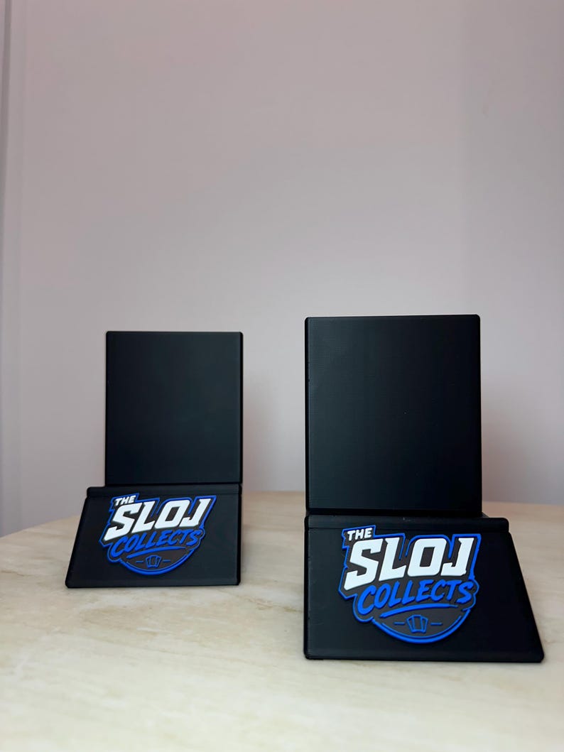 May include: Two black display stands with the text "THE SLOJ COLLECTS" in white and blue. The stands have a rectangular base and a square top. The stands are on a light-colored surface.